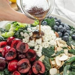 Dressing being poured on top of a summer kale salad with fresh cherries