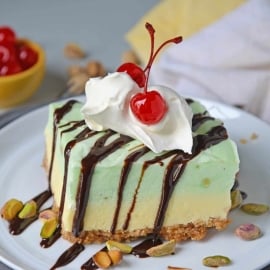 This no-bake cake is layered with graham crackers, vanilla and pistachio pudding and topped with chopped pistachios, chocolate sauce and cherries! #iceboxcake #pistachioiceboxcake #pistachiocake www.savoryexperiments.com