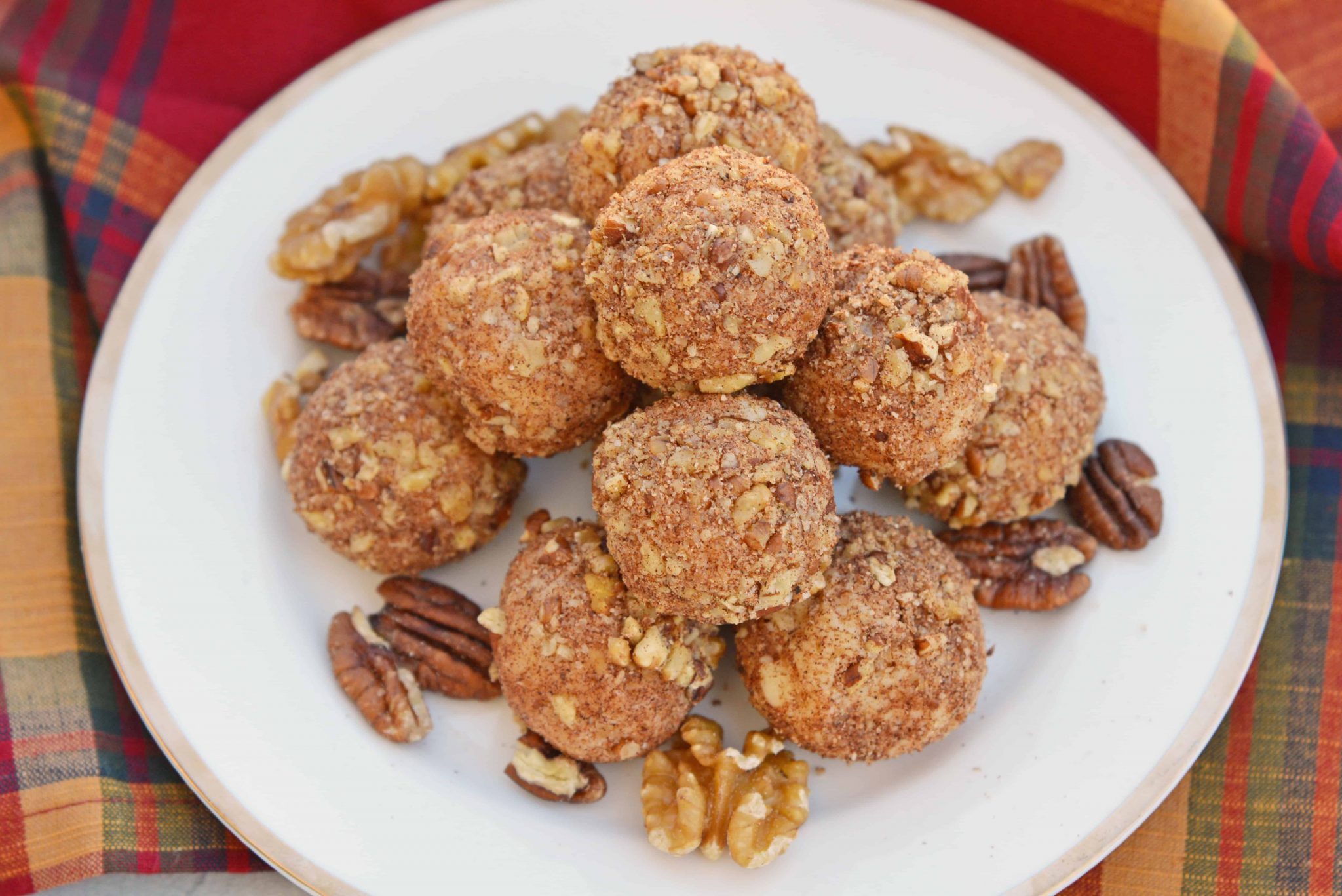 A white plate is filled with a stack of round cookies coated in a crumbly mixture of nuts and spices. The plate is garnished with whole walnuts and pecans. A colorful plaid cloth is partly visible in the background.