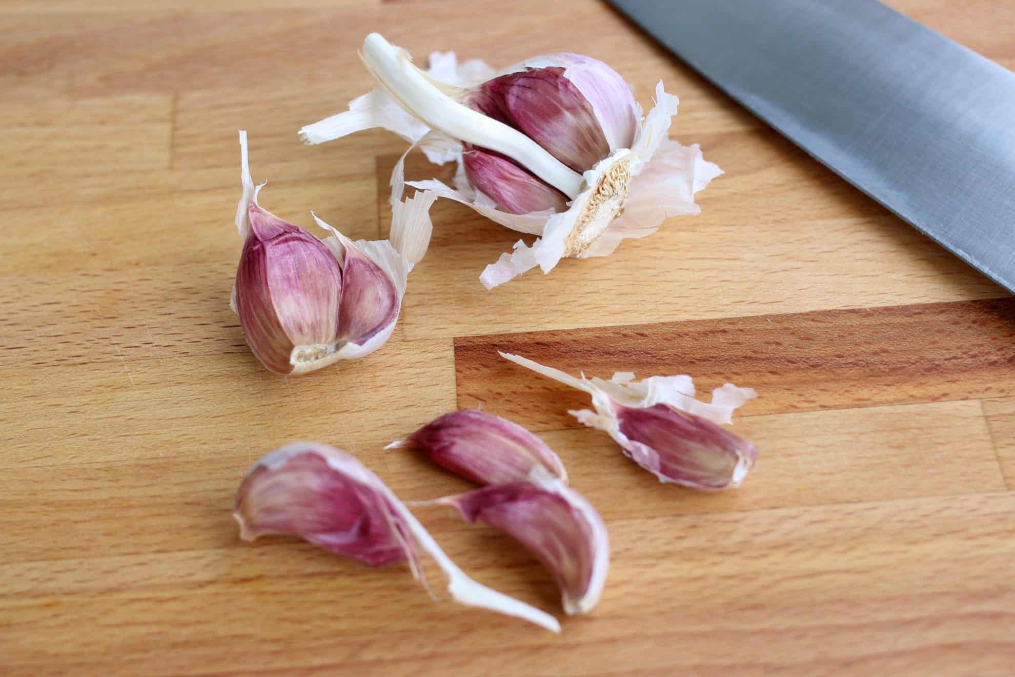 Whole garlic cloves setting on a wooden cutting board alondside a chefs' knife.