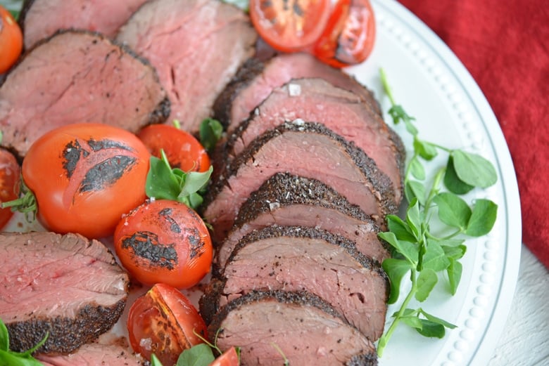 close up of sliced coffee crusted beef tenderloin with pea shoots and tomatoes