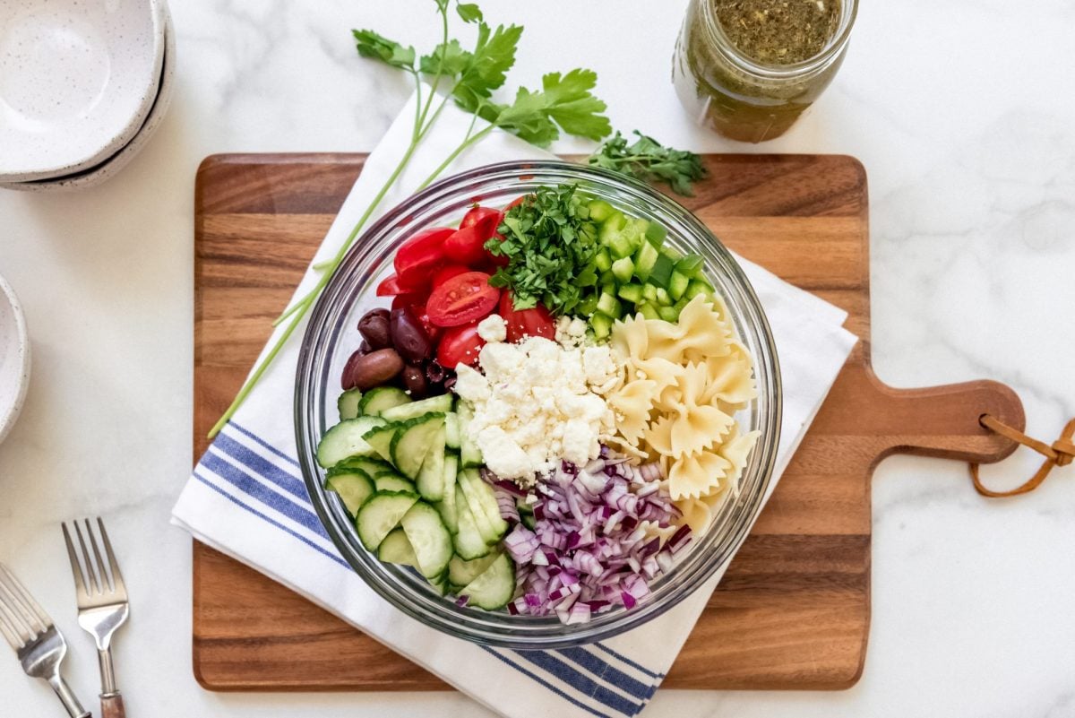 ingredients for Greek pasta salad in a large mixing bowl on a wood cutting board 