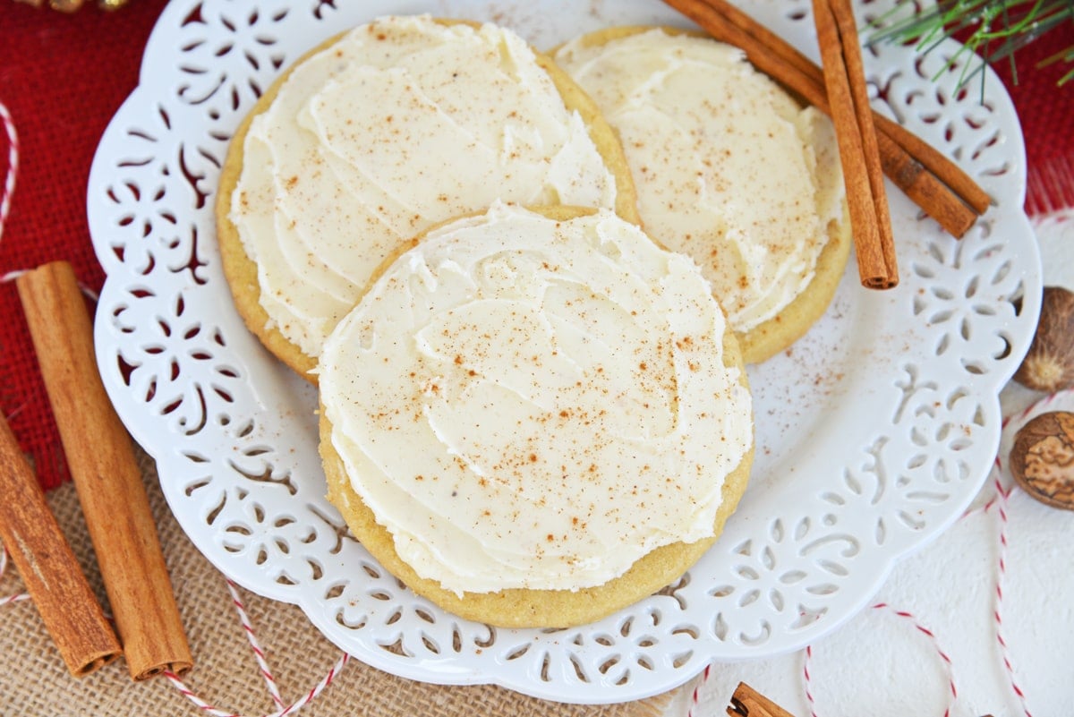 three frosted eggnog cookies on a white serving plate with cinnamon sticks