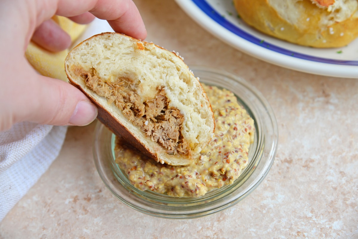 hand dipping pulled pork stuffed pretzel roll into mustard