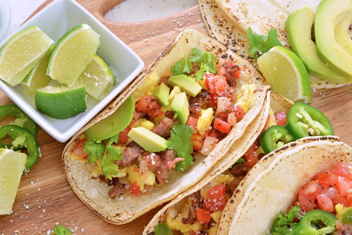 close up of an eggs cheese avocado taco with limes on a wood board