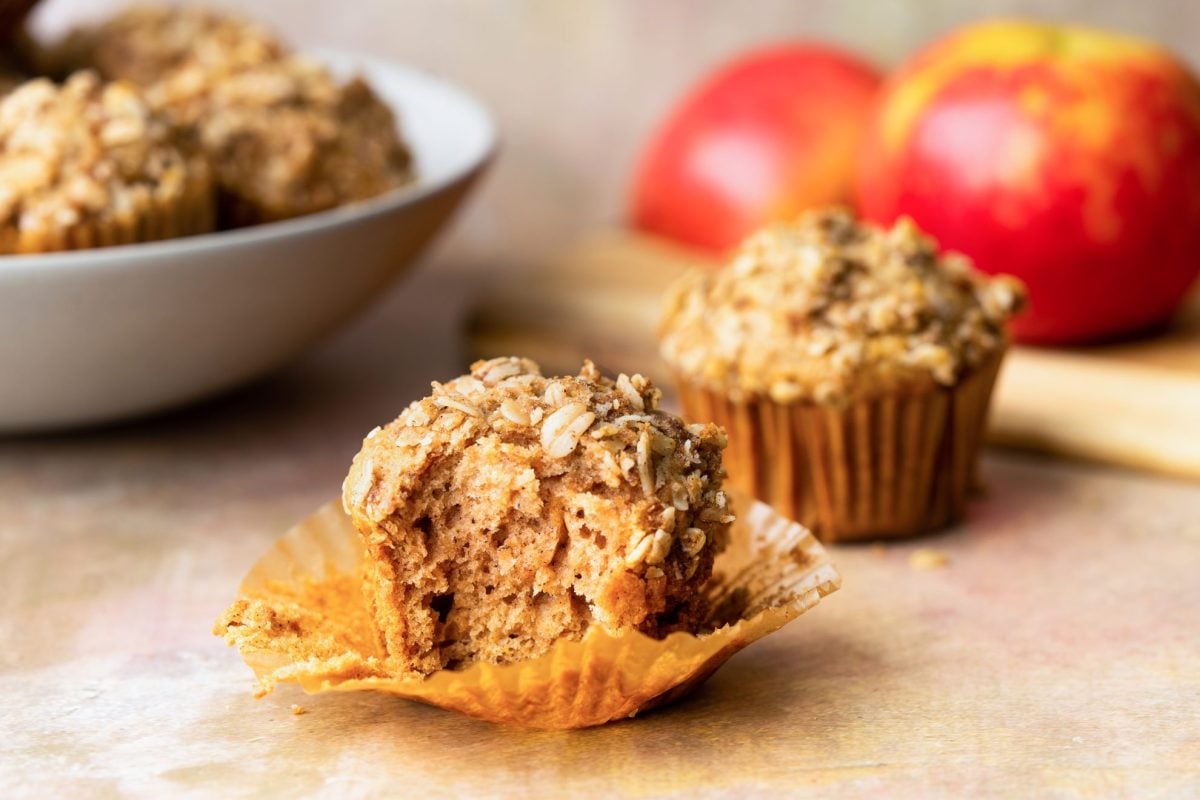 Applesauce muffin with bite taken out of it and a bowl of muffins and apples in the background