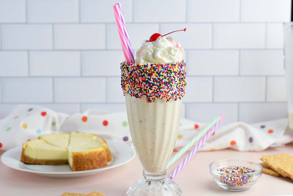 head-on image of a cheesecake milkshake with cheesecake in the background and colorful straws 