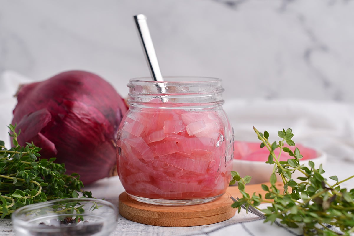 head on view of pickled onions in a small glass jar with a fork and fresh herbs 