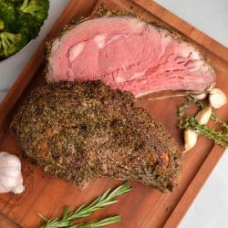 overhead of rare rib roast on a cutting board with fresh herbs and garlic cloves