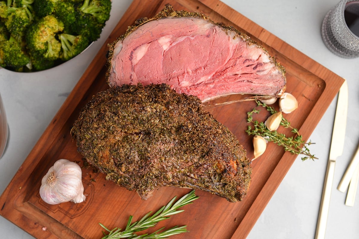 overhead of rare rib roast on a cutting board with fresh herbs and garlic cloves