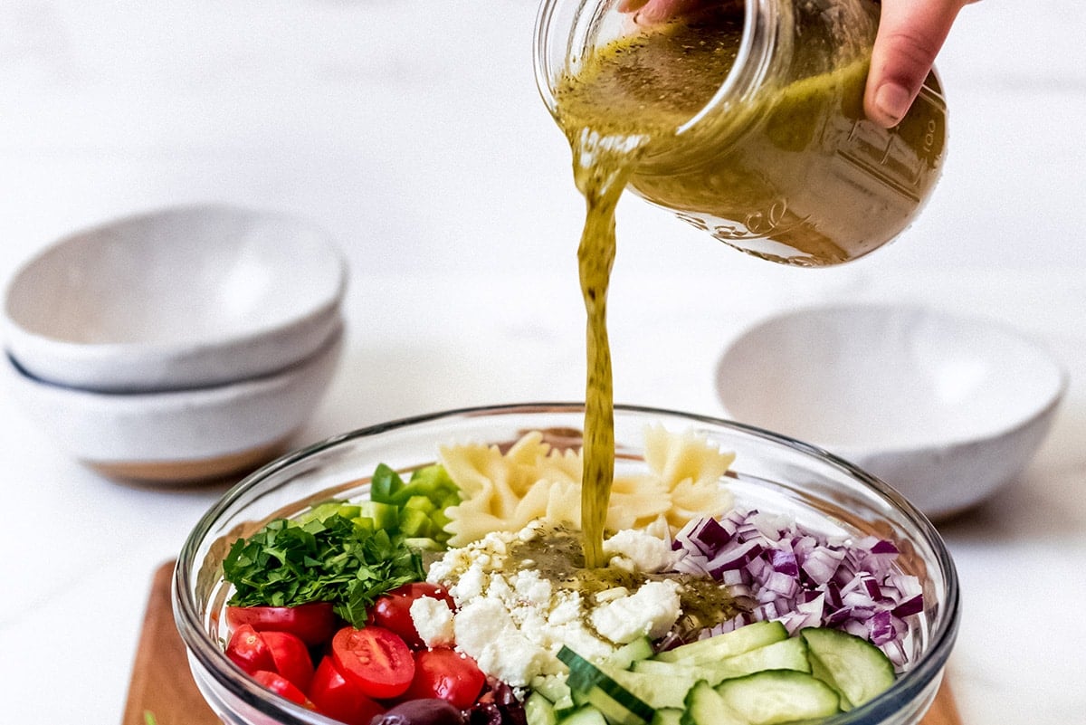 pouring greek salad dressing over pasta salad 