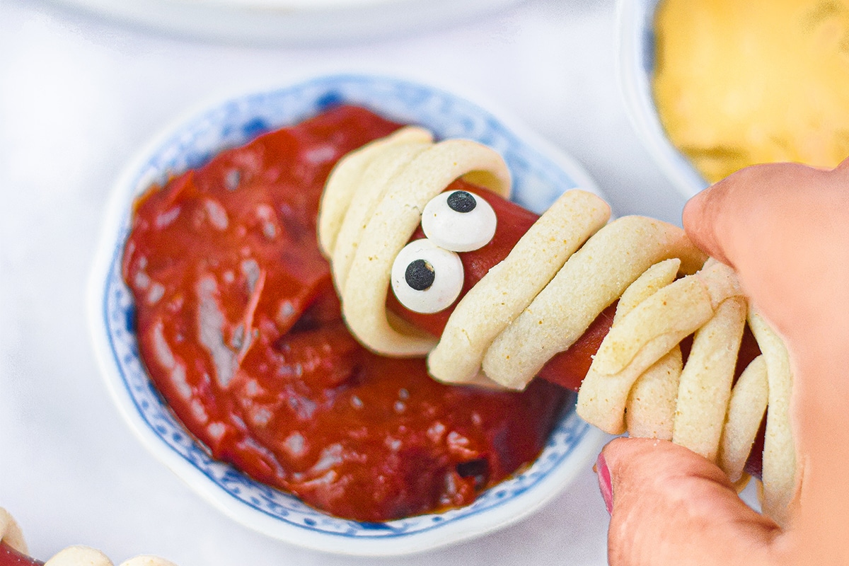 hand dipping mummy hot dog in ketchup