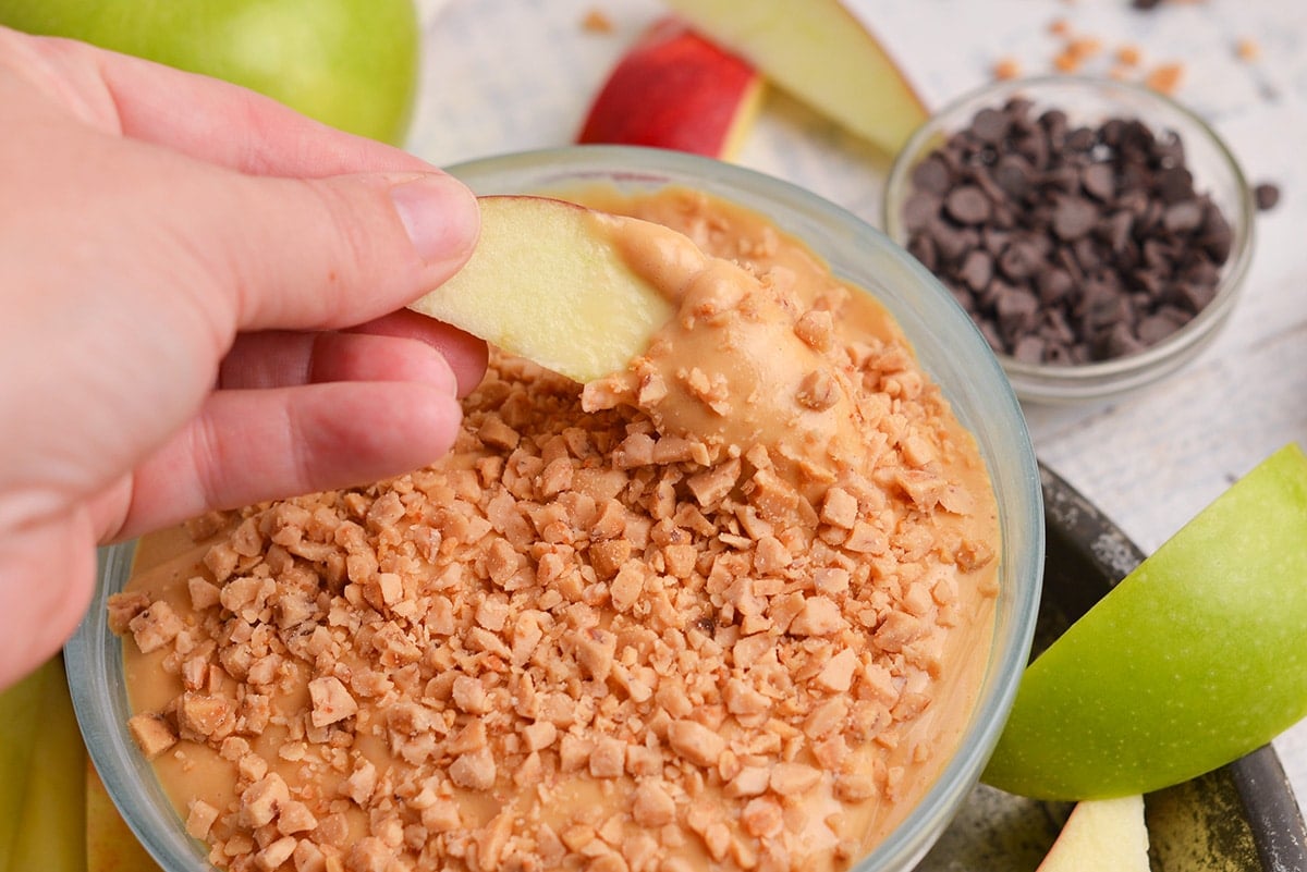 hand dipping apple slice into caramel apple dip with toffee pieces