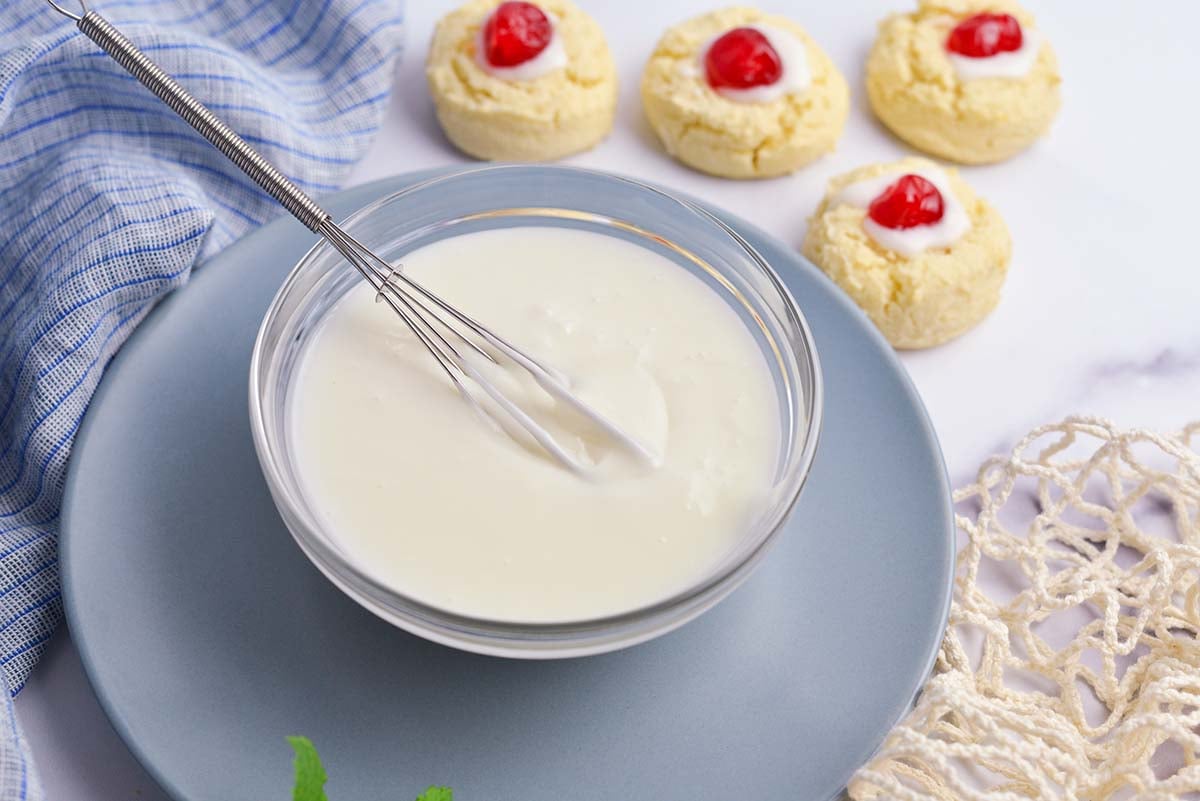 small whisk in a bowl of glaze on a blue plate with cookies in the background