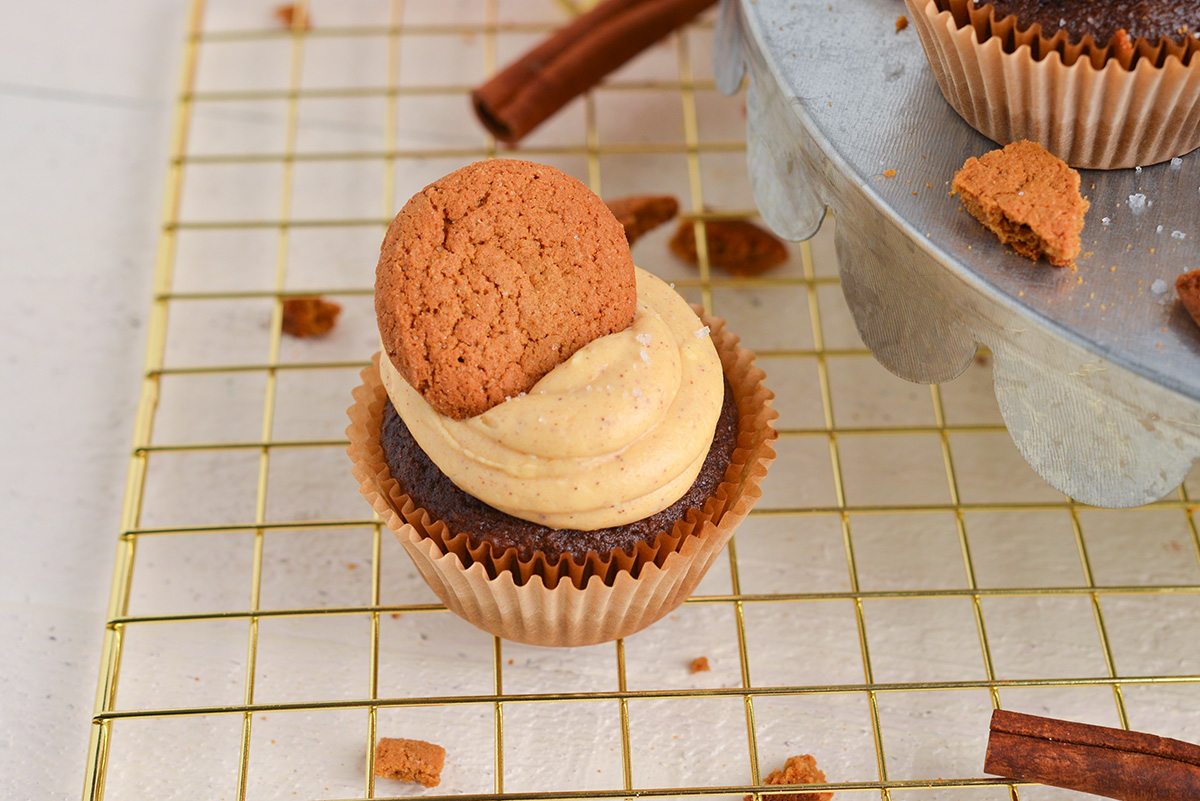 angled shot of chocolate cupcake on cooling rack topped with pumpkin cream cheese frosting and a cookie