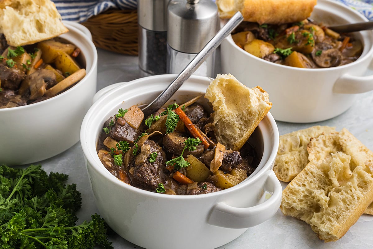angled shot of spoon in crock of red wine beef stew with crusty bread
