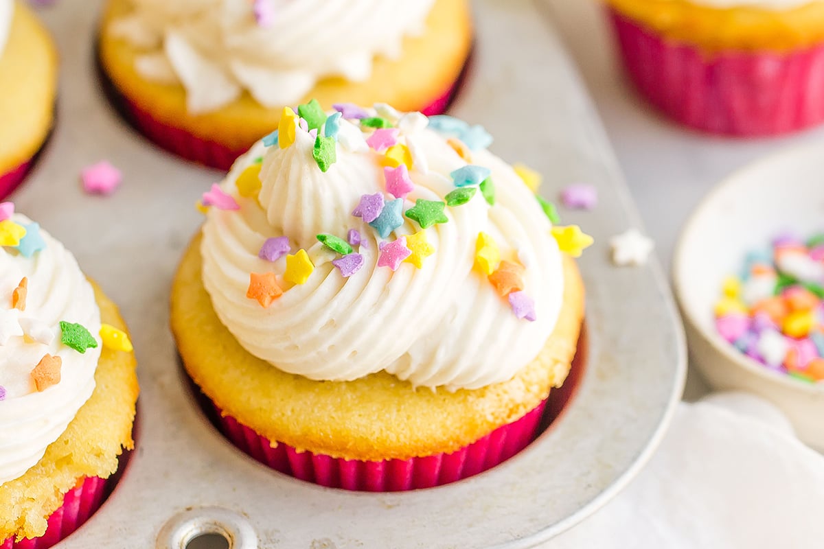 close up of vanilla cupcakes with frosting and sprinkles in the cupcake tin