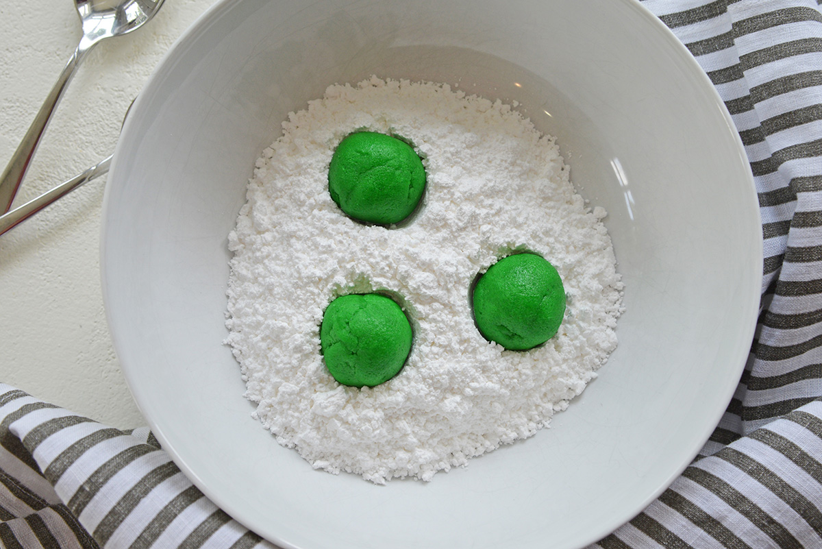 three green balls of cookie dough in bowl of powdered sugar
