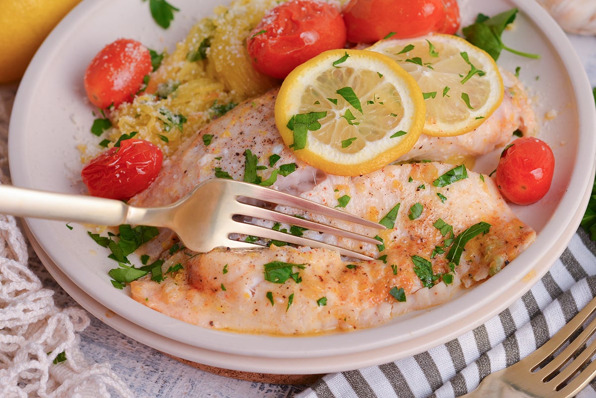 angled shot of fork digging into baked tilapia on plate