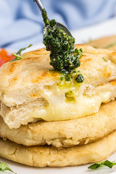 spoon adding chimichurri sauce to stack of arepas