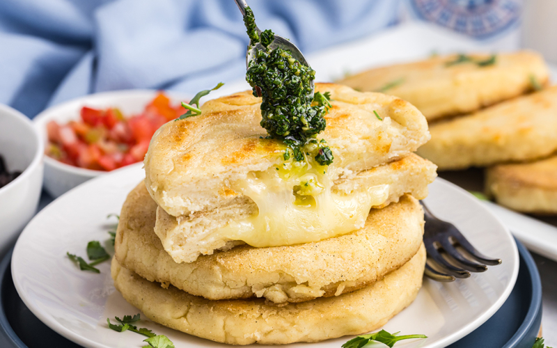 spoon adding chimichurri sauce to stack of arepas
