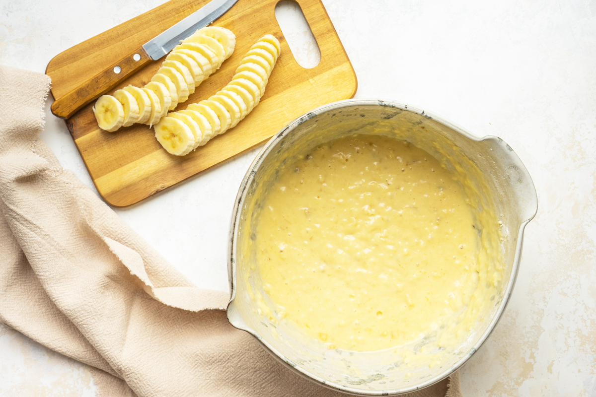 overhead shot of pancake batter in bowl with sliced bananas on cutting board