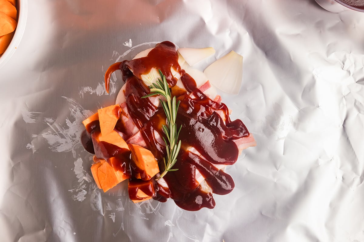 overhead shot of rosemary sprig and raw sweet potatoes added to chicken foil packets