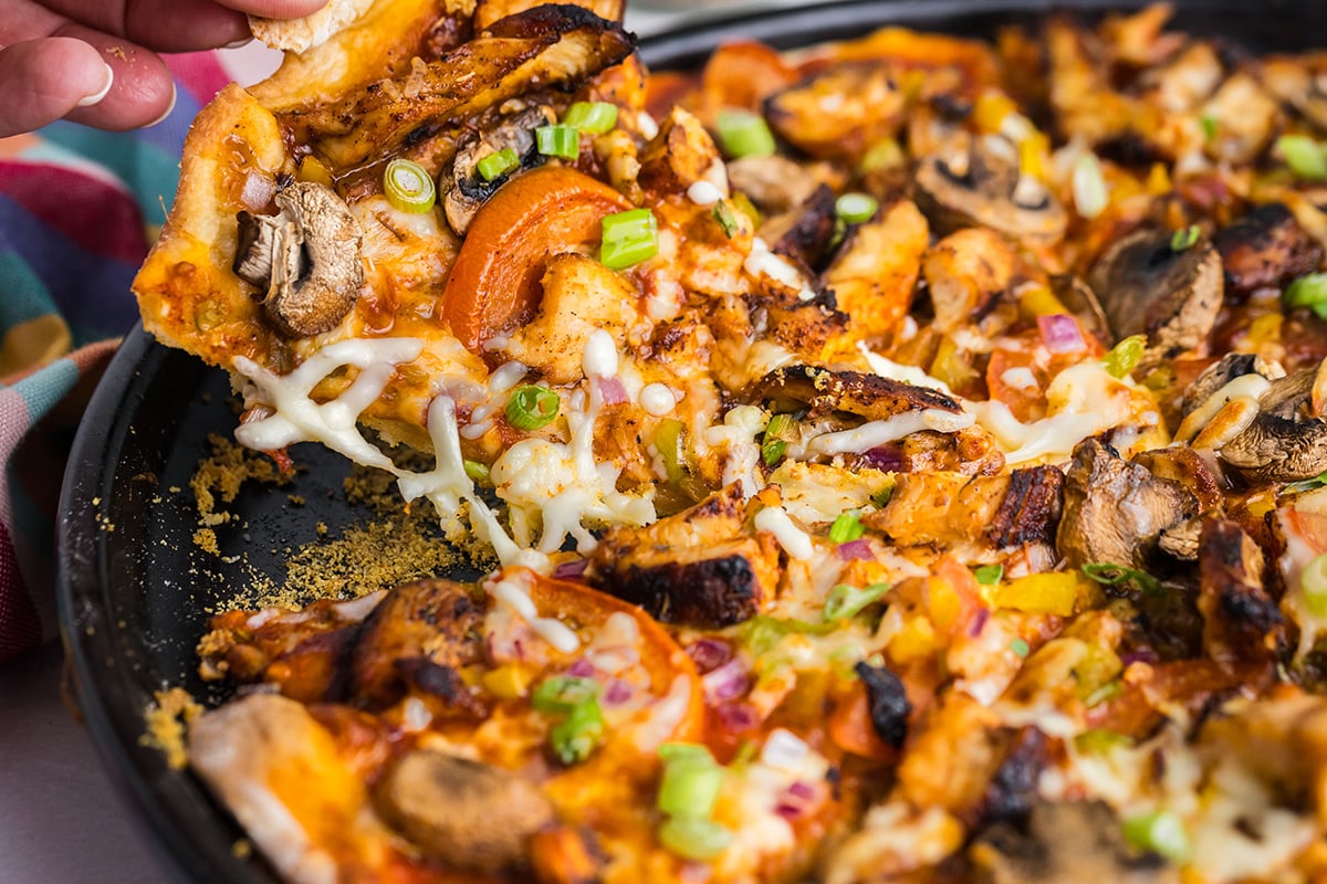 angled shot of hand grabbing slice of jerk chicken pizza