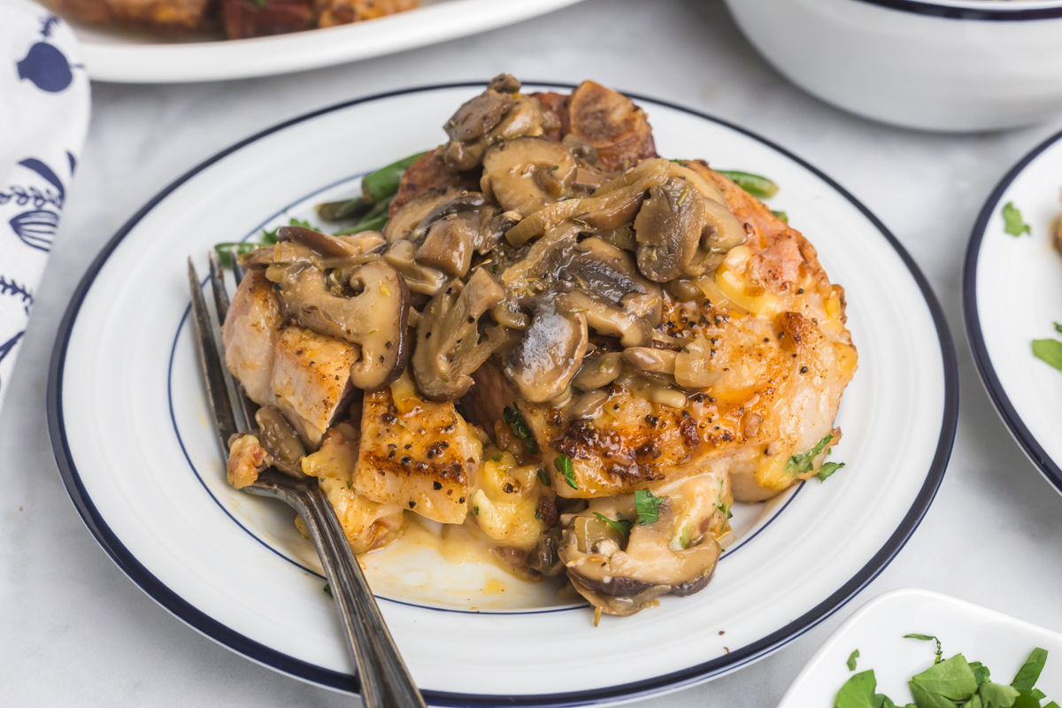 close up of baked stuffed pork chop topped with mushrooms on plate with fork