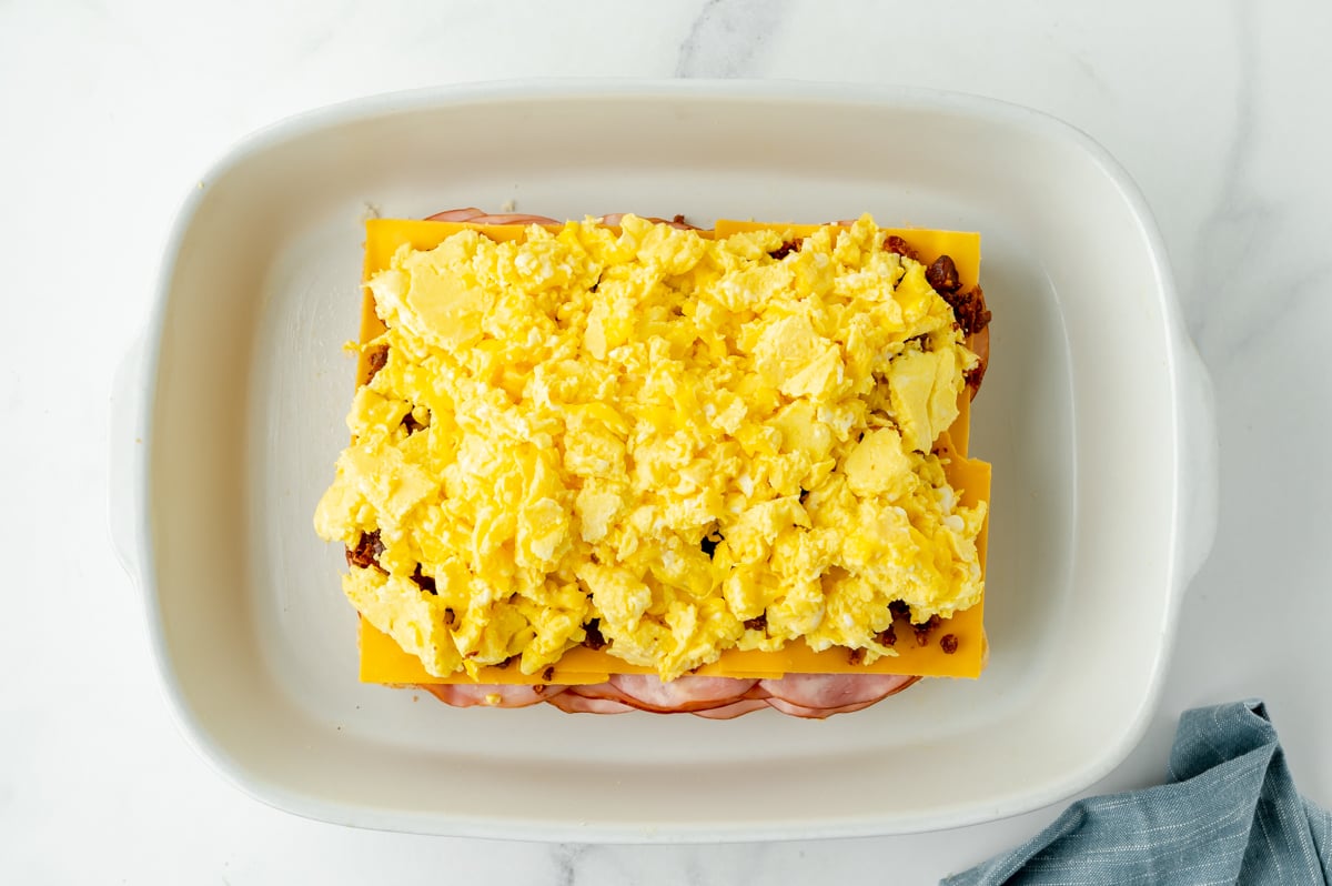 overhead shot of scrambled eggs over cheese, bacon and ham on bottom slider buns