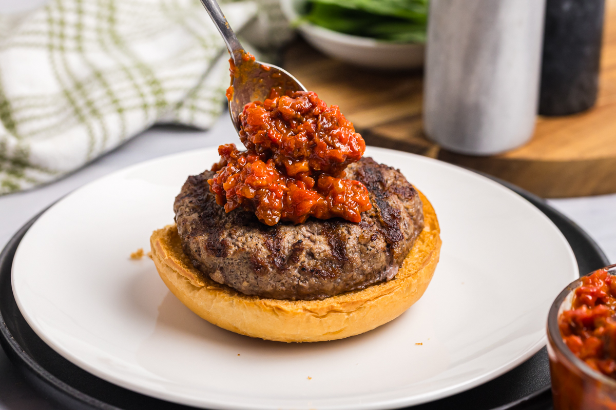 Spoonful of sun-dried tomato and roasted red pepper spread being added to a grilled caprese burger