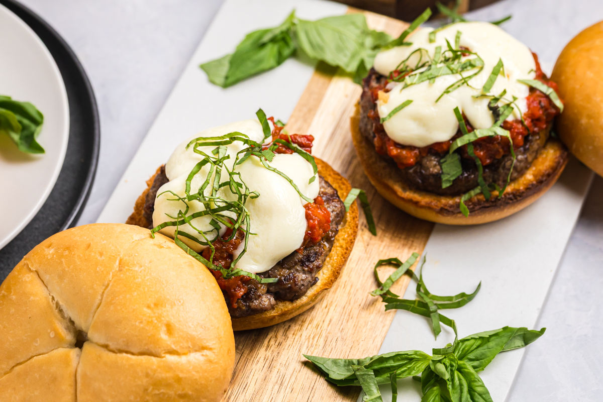 Two grilled caprese burgers topped with mozzarella and basil on a wooden serving board